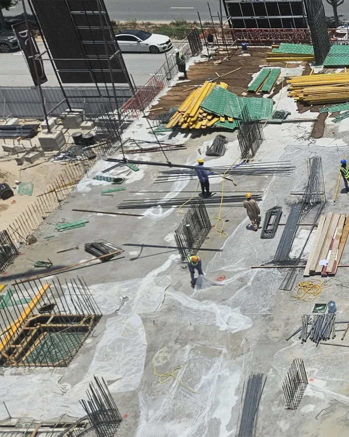 Construction site with workers in hard hats laying out steel rebar and stacked yellow/green panels on a concrete slab. Crates, hoses, and scattered tools are visible across the area.