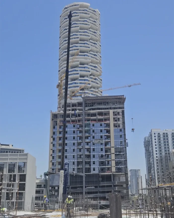 Under-construction high-rise with a twisting, curved façade and a crane nearby; construction workers on the ground level.