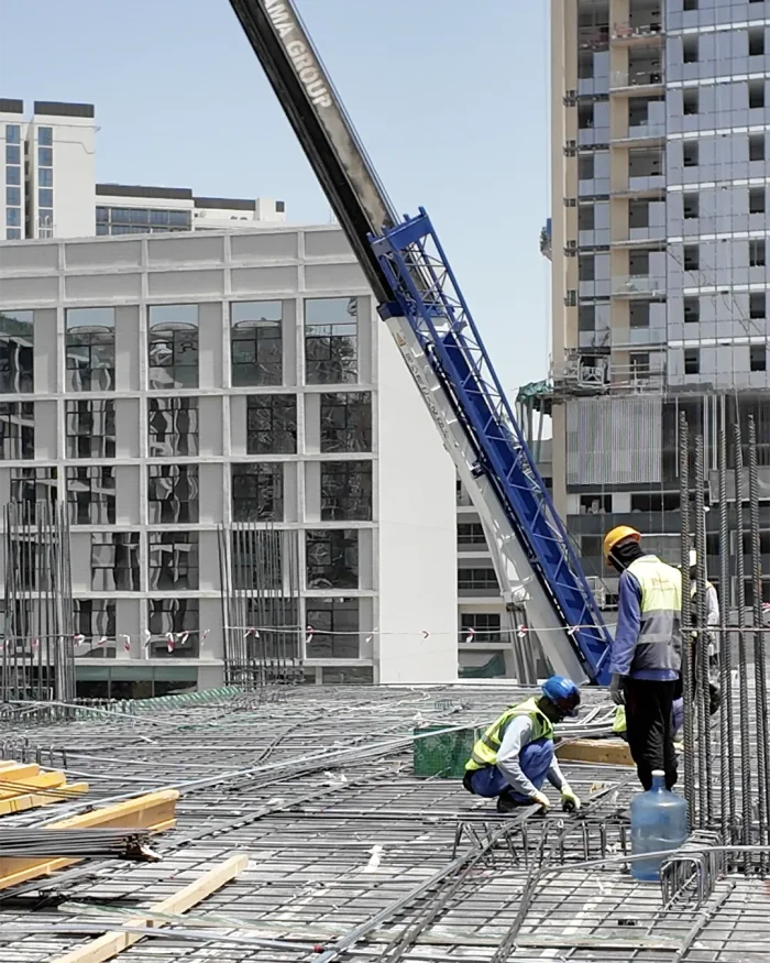 Construction workers in high-visibility vests and hard hats tie steel rebar on a concrete slab at a high-rise construction site, with a blue crane arm overhead and glass buildings in the background,