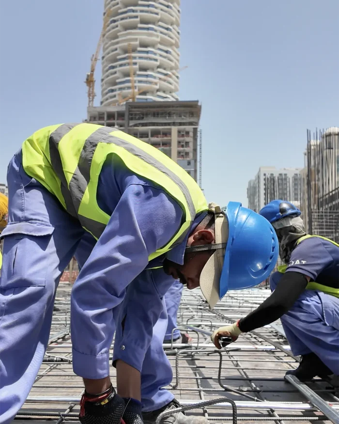 Workers in blue uniforms and lime-yellow vests bending over rebar on a construction site with a tall building under construction in the background.