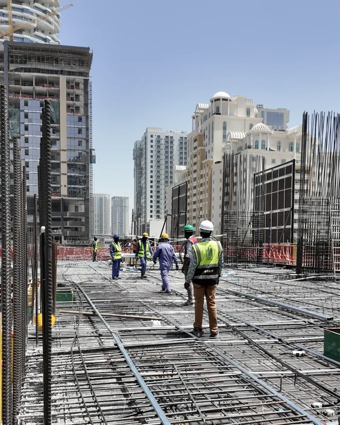 Construction workers in safety gear standing on a steel rebar grid at a high-rise construction site.