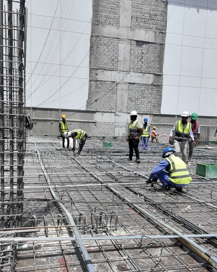 Construction workers in high-visibility vests and hard hats lay and inspect steel rebar for a concrete slab.