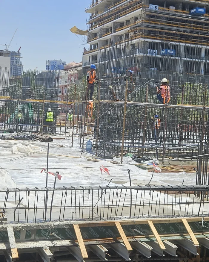 Construction site with workers in safety gear and helmets on scaffolding around a reinforced concrete framework under construction.