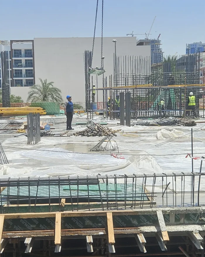 Construction site with steel rebar, concrete slabs, and a crane lifting a metal plate above workers wearing hard hats and vests on a sunny day.