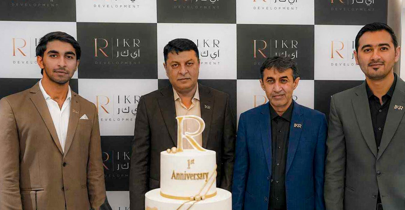 Four men in suits stand beside a two-tier white and gold anniversary cake, with a large gold 'R' topper, in front of a checkered backdrop featuring 'IKR' logos.